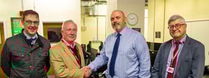 Four men standing in a straight line facing the camera, with the central pair shaking hands